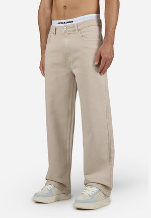 Beige weite Jeans mit einem kontrastierenden weißen Bund, der mit dem 'PEGADOR'-Branding versehen ist. Getragen mit hellen Sneakers.