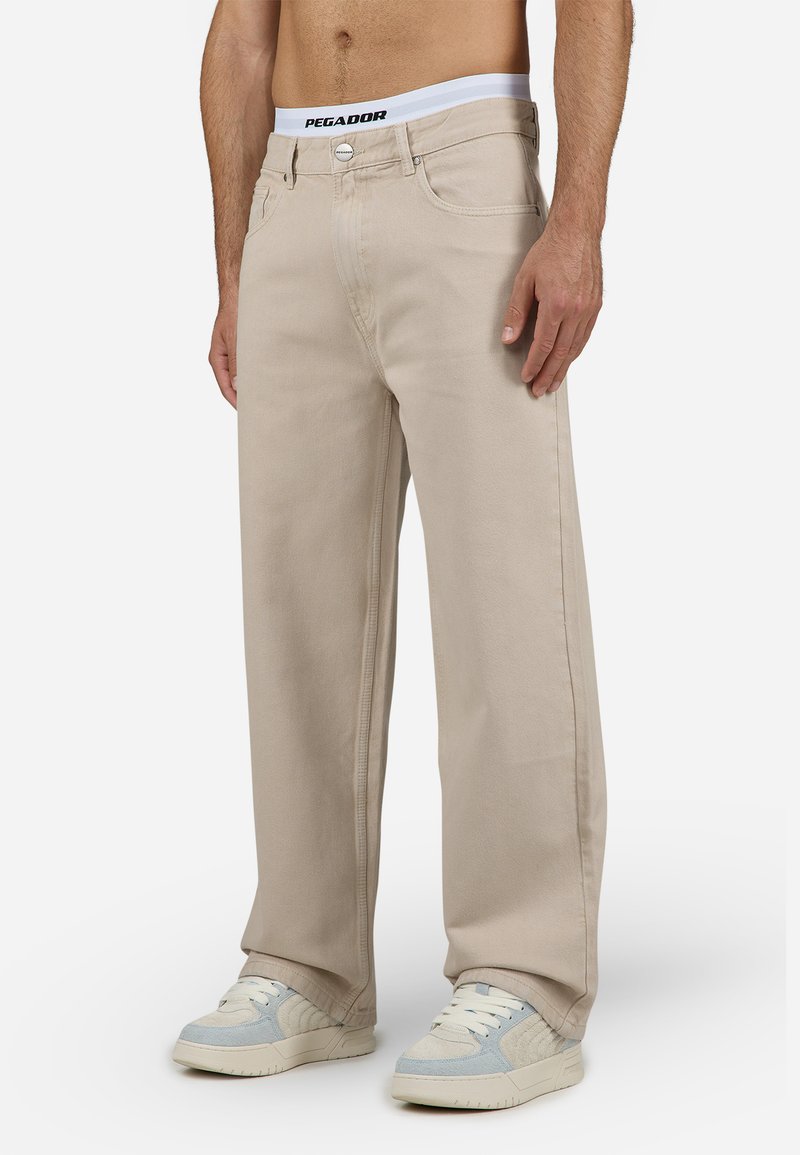 Beige wide-leg jeans with a contrasting white waistband featuring 'PEGADOR' branding. Worn with light-colored sneakers.