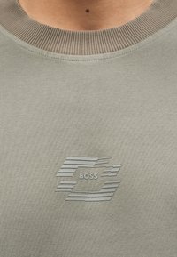 Κοντινή λήψη γκρι μπλούζας με στρογγυλή λαιμόκοψη και ριμπάτο περιθώριο, με στυλιζαρισμένο λογότυπο "BOSS" στο κέντρο.