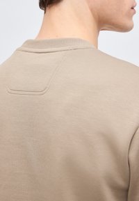 Μπεζ τοπ με στρογγυλή λαιμόκοψη που δείχνει την περιοχή των ώμων στο πίσω μέρος με ορατά ραφτά και κοντό λαιμό που φαίνεται με φόντο μια απλή επιφάνεια.