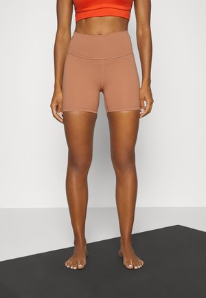 Mujer de pie descalza sobre una alfombrilla negra, vistiendo unos pantalones cortos marrones de talle alto y un top corto naranja, mostrada desde el torso medio hasta los pies.