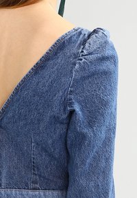 Denimmekko syvällä V-pääntiehellä, puffihihat ja tiukka vartalo. Kangas on sininen ja siinä on tekstuuripinta, jossa on ompeluyksityiskohtia.