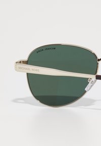 Lunettes de soleil en métal doré avec des verres verts. La monture présente une forme arrondie, des branches fines, et "MICHAEL KORS" gravé sur le côté.