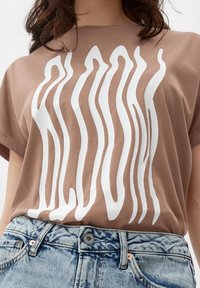 Hnědé bavlněné tričko s bílým vlnitým vzorem na přední straně. Kombinováno s světle modrými džínovými šortkami s klasickým zapínáním na knoflíky.