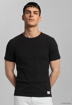 Νέος άνδρας με κοντά σκούρα μαλλιά φοράει μαύρο απλό κοντομάνικο μπλουζάκι και λευκό παντελόνι, στέκεται μπροστά σε ανοιχτό γκρι φόντο.