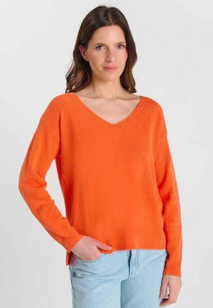 Vrouw met bruin haar die een feloranje V-hals trui en lichtblauwe spijkerbroek draagt, poserend met één hand in haar zak tegen een effen achtergrond.