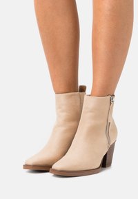 Beige suede ankle boots with a pointed toe, side zipper, and a stacked brown heel. Smooth texture and minimalist design.