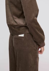 Barna kordbársony melegítő, amely függőleges bordázattal, bordázott mandzsettákkal és oldalzseb részlettel rendelkezik. Puha tapintású és kényelmes szabású.