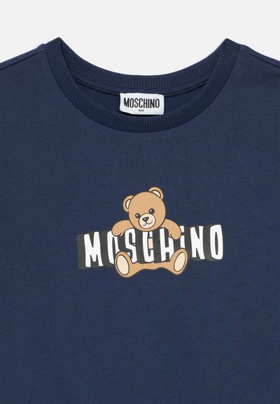 Παιδικό πουκάμισο σε σκούρο μπλε με ριμπ γιακά, διακοσμημένο με γραφικό αρκουδάκι που αγκαλιάζει το κείμενο "Moschino" σε λευκά και μαύρα γράμματα τύπου block.