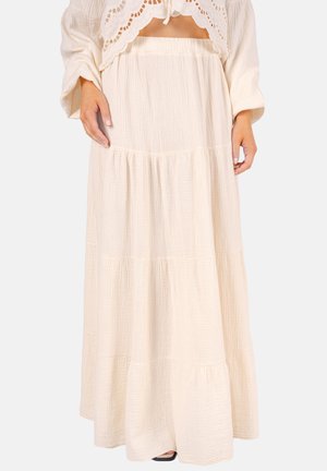 Frau trägt einen cremefarbenen, gestuften Maxirock aus strukturiertem Stoff mit elastischem Bund, kombiniert mit einem passenden langärmligen Oberteil.