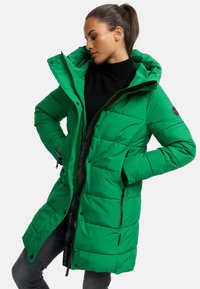 Abrigo acolchado verde con capucha, que cuenta con un cierre frontal, bolsillos laterales y un diseño acolchado. Tela texturizada y logotipos visibles en las cremalleras.