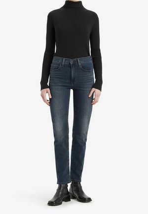 Personne portant un pull à col roulé côtelé noir, un jean taille haute bleu foncé et des bottines noires, debout sur fond blanc.