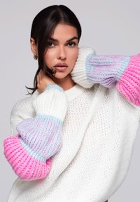 Pull en tricot blanc avec des manches surdimensionnées à motifs roses et bleus. Le fil texturé apporte de la dimension aux manches colorées.