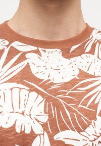 Κοντινό πλάνο του λαιμού και του άνω μέρους του στήθους ενός ατόμου που φοράει καφέ πουκάμισο με λευκό τροπικό λουλουδένιο σχέδιο.