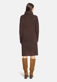 Femme de dos portant une robe-pull longue marron à col roulé avec manches longues et des bottes en daim beige montant jusqu'aux genoux.