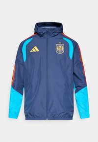 Tmavomodrá bunda s kapucňou, s modrými rukávmi, červenými a žltými akcentmi a zlatým logom Adidas. Na prednej strane je znak národného tímu Španielska.
