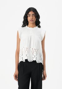 Blouse en coton blanc avec des manches froncées, présentant des découpes brodées en bas dans un motif floral. Assortie avec un pantalon noir ample.