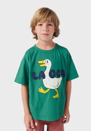 Grünes Baumwoll-T-Shirt mit einem Cartoon-Enten-Motiv in Weiß und Gelb. Text "LA OCA" in Dunkelblau unter der Abbildung. Kurze Ärmel.