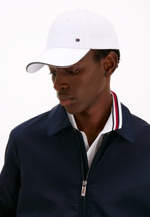 Casquette de baseball blanche avec une visière courbée et un petit logo. Portée avec une veste bleu marine présentant un col rayé et une fermeture éclair.
