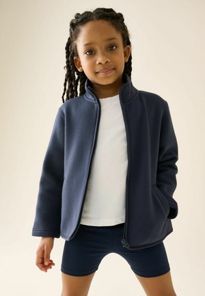 Jeune enfant aux cheveux tressés portant une veste zippée bleu marine, une chemise blanche et un short bleu marine, debout devant un fond clair.