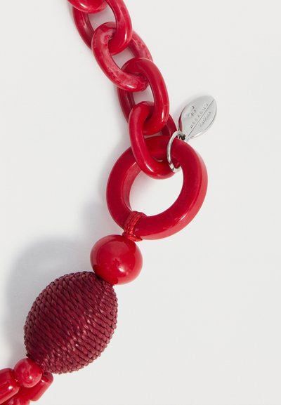 Κόκκινες πλαστικές αλυσίδες συνδεδεμένες με κόκκινη υφασμάτινη οβάλ χάντρα, στρογγυλή χάντρα και ασημένια ετικέτα με χαραγμένο λογότυπο σε λευκό φόντο.