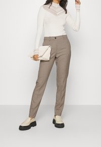 Vrouw in een beige top met kantafwerking en lange mouwen, bruine geblokte broek, beige platform instappers, met een beige clutch tas.