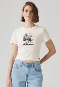 Kortärmad, krämfärgad t-shirt i bomull med en svartvit basset hound-grafik och text, croppad design. Ljusblå jeans bärs.