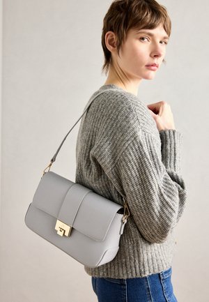 Person trägt einen grauen Strickpullover und blaue Jeans, trägt eine graue Schultertasche mit goldener Schnalle über der Schulter und schaut seitlich.