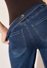 Jeans de mezclilla azul oscuro con bolsillos traseros con costura superior y una etiqueta de marca negra visible a lo largo de la cinturilla.