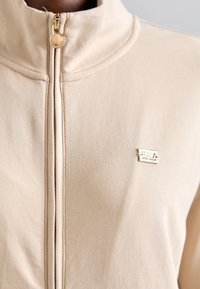 Béžová bunda na zips s vysokým golierom, hladkou textúrou a zlatým logom na ľavej hrudi. Pritiahovač zipsu má okrúhly tvar.