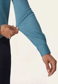 Lichtblauw shirt met lange mouwen en een gladde textuur, voorzien van een aansluitende manchette en rekbare stof; hand die voorzichtig aan de mouw trekt.
