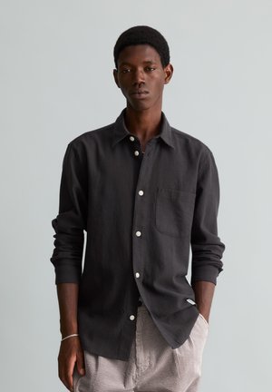 Jeune homme portant une chemise sombre à manches longues boutonnée et un pantalon en velours clair, debout avec une main dans la poche sur un fond uni.
