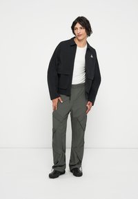 Casaco preto com fecho éclair e dois bolsos no peito, usado sobre uma camisola branca. Calças cargo verde oliva com detalhes em zíper e bolsos. Calçado preto.