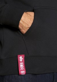 Sudadera negra con capucha, puños acanalados y un bolsillo frontal, con una etiqueta de tela roja prominente con texto blanco en la costura lateral.