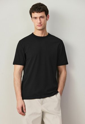 Young man with short dark hair wearing a plain black short-sleeve t-shirt and light beige pants, standing against a light gray wall.