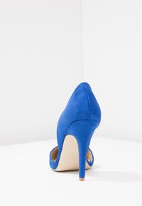 Chaussure à talon haut en daim bleu avec un bout pointu et un talon fin de couleur claire. Le design présente une texture lisse et une silhouette épurée.