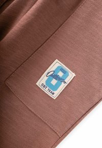 Hnedá tkanina s hladkým povrchom, na boku švu s modro-bielou nálepkou s nápisom "2010 Outfitters 6 COT TEAM."