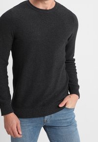 Dunkelgrauer Strickpullover mit Rundhalsausschnitt, strukturiertem Stoff, gerippten Bündchen und Saum. Getragen mit hellblauen Jeans, die Hände in den Taschen.