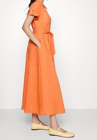 Maxi šaty z oranžového lnu s krátkými rukávy, páskem v oblasti pasu, dvěma bočními kapsami a límečkem. Oblečené s žlutými plochými botami.