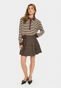 Woman wearing brown and white striped long-sleeve polo shirt, brown pleated skirt with blue accents, and black pointed slingback shoes with bows.