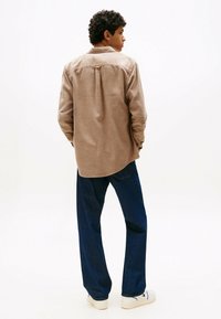 Brown button-up shirt with a smooth texture, visible stitching, and a yoke design, paired with dark blue wide-leg jeans and white sneakers.