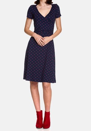 Nainen pukeutuneena tummansiniseen polvipituiseen mekkoon, jossa on punaisia sydänkuvioita, ja punaisiin nilkkasaappaisiin, seisoo kädet edessä ristiin asetettuina.