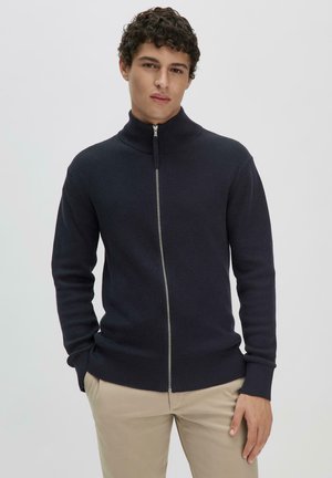 Marineblauer, gerippter Strick-zipper mit hohem Kragen, langen Ärmeln und kontrastierendem silbernem Reißverschluss. Getragen mit hellfarbigen Hosen.