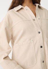 Beige denim jacket with a collar, snap buttons, and two front pockets. Features a textured fabric with subtle ribbing and metal accents.