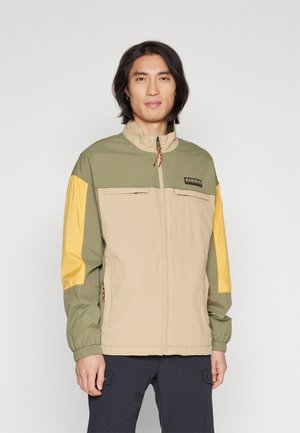 Chaqueta beige y verde con mangas amarillas, cierre de cremallera y dos bolsillos frontales. Cuenta con un alto cuello y cordón con estampado.