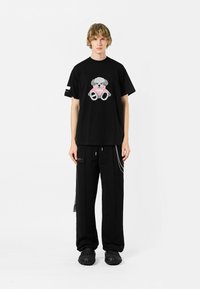 Camiseta negra de algodón con un gráfico texturizado de un oso gris con acentos en rosa, combinada con pantalones holgados negros y calzado negro grueso.