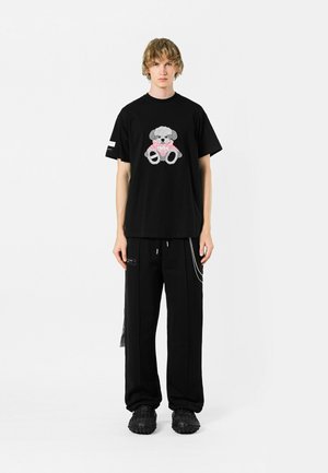 Camiseta negra de algodón con un gráfico texturizado de un oso gris con acentos en rosa, combinada con pantalones holgados negros y calzado negro grueso.