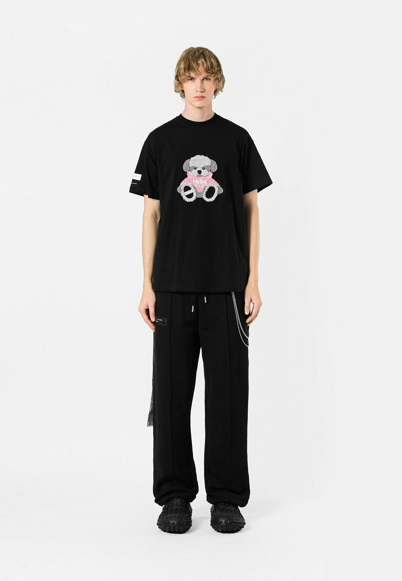 Camiseta negra de algodón con un gráfico texturizado de un oso gris con acentos en rosa, combinada con pantalones holgados negros y calzado negro grueso.
