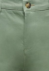 Détail avant d'un pantalon vert montrant la fermeture par bouton, la ceinture et la texture lisse du tissu.