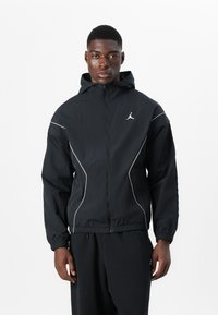 Veste coupe-vent noire avec capuche, dotée d'accents blancs et d'un logo Jordan sur la poitrine. Poignets et taille élastiques. Matériau léger.